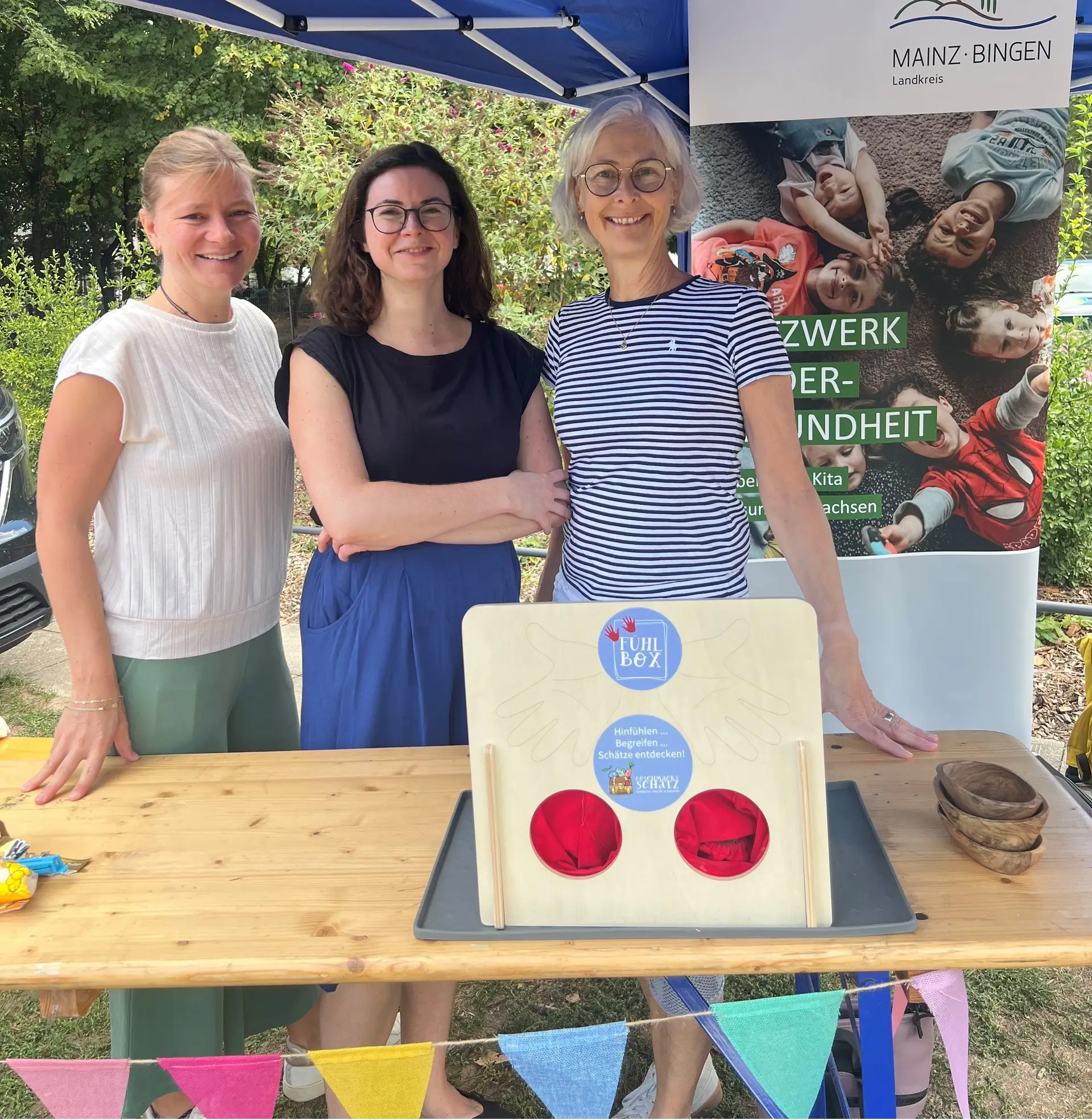 Drei Frauen stehen nebeneinander und lächeln in einer Kinderveranstaltung. Im Vordergrund sind Banner oder Infomaterial des Präventionsteams zu sehen.