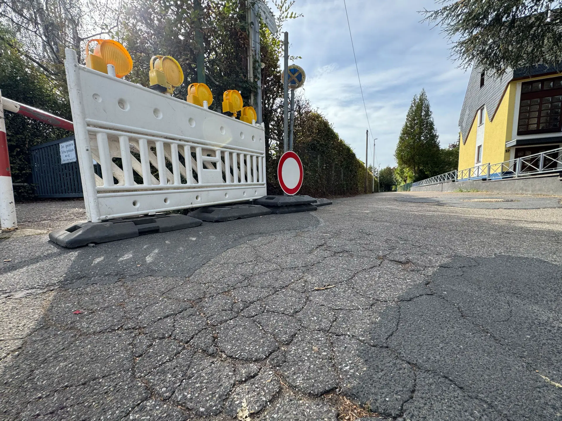 Eine weiße Baustellenabsperrung mit gelben Warnleuchten steht auf rissigem Asphalt vor der Zufahrt zur Grundschule Kesselheim.